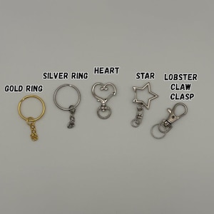 May include: A collection of metal keychain hardware including a gold ring, a silver ring, a heart-shaped clasp, a star-shaped clasp, and a lobster claw clasp. Each item is labeled with its corresponding name in black text above the hardware.