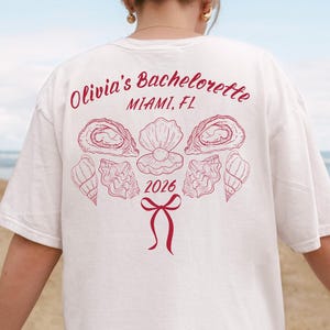 以下が含まれることがあります： 白いTシャツ、赤いデザイン。「Olivia's Bachelorette Miami, FL 2026」の文字と貝殻とリボンのイラスト。カジュアルなスタイルのTシャツです。