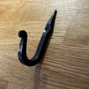May include: A hand-forged, black metal hook with a curved design. The hook has a rounded end and a pointed tip with a small hole. The hook is designed to be mounted on a wall or other surface for hanging items.