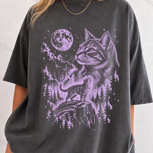 May include: A charcoal grey short-sleeve t-shirt featuring a light purple graphic print. The design displays a large cat head, a smaller cat standing on a rocky cliff, a full moon, and a forest of pine trees against a starry night sky.