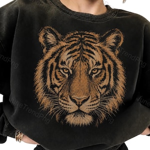 Puede incluir: Una sudadera negra con efecto lavado al ácido que presenta un gran estampado gráfico detallado de la cara de un tigre en tonos marrones y naranjas. La sudadera tiene cuello redondo, mangas largas con puños acanalados y un corte holgado.