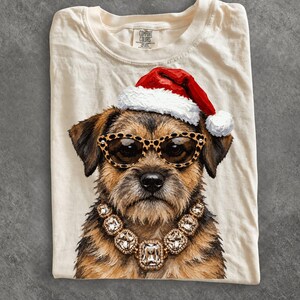 Könnte beinhalten: Ein cremefarbenes T-Shirt mit einer Grafik eines braunen Terrier-Hundes, der eine rot-weiße Weihnachtsmütze, eine Sonnenbrille mit Leopardenmuster und eine große, klobige goldfarbene Halskette mit klaren rechteckigen Kristallen trägt.