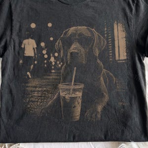 May include: A charcoal grey t-shirt featuring a tan-coloured graphic print of a dog wearing sunglasses. The dog is positioned behind a clear plastic cup with a straw. The background of the print shows a blurry street scene with circular light orbs.
