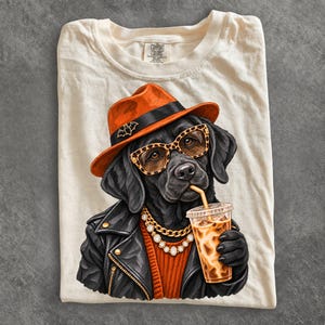 May include: A cream-colored t-shirt featuring a graphic of a black Labrador dog wearing an orange fedora with a bat pin, leopard print glasses, a gold chain necklace, and a black leather jacket while holding an iced coffee in a plastic cup.