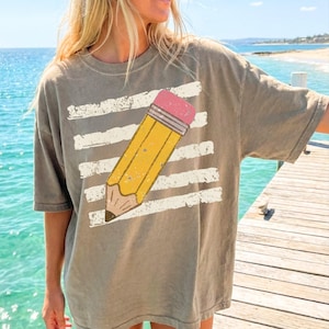 Puede incluir: Camiseta verde oliva de manga corta con un diseño de lápiz amarillo y goma rosa. El lápiz está sobre rayas horizontales blancas, dando un aspecto envejecido. La camiseta es de estilo casual y cómodo.