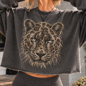 May include: A charcoal gray cropped sweatshirt featuring a detailed graphic print of a leopard face in shades of brown, tan, and black. The garment has a crew neckline, long sleeves, and a raw, unfinished hemline at the bottom edge.