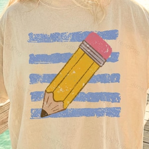 Puede incluir: Camiseta color crema con un gran dibujo de un lápiz amarillo, una goma de borrar rosa y rayas horizontales azules. El lápiz está inclinado sobre la camiseta, creando un diseño lúdico. La camiseta tiene un aspecto vintage y desgastado.