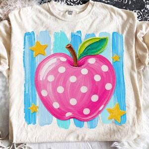 Peut inclure: Un t-shirt couleur crème arborant un motif de pomme rose à pois blancs, avec une tige brune et une feuille verte. La pomme est centrée sur des coups de pinceau bleu clair verticaux et entourée de quatre étoiles jaunes dans les coins.