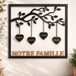 Pode incluir: Uma peça de decoração de parede quadrada com moldura preta e fundo branco. Um ramo de árvore preto estende-se pelo topo com quatro ornamentos pretos em forma de coração pendurados, cada um com um nome. O texto NOTRE FAMILLE está na base.