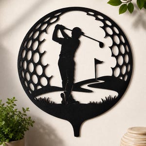 Könnte beinhalten: Ein kreisförmiges Wandkunstobjekt aus schwarzem Metall mit der ausgeschnittenen Silhouette eines Golfers im Schwung. Das Design ist von einem Muster umrahmt, das an die Noppenoberfläche eines Golfballs erinnert, mit Flagge und Grün.
