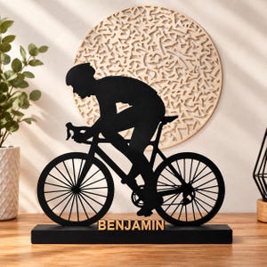 Könnte beinhalten: Eine schwarze Metallsilhouette eines Radfahrers auf einem Rennrad, montiert auf einem rechteckigen schwarzen Sockel. Der Name BENJAMIN ist in goldenen Großbuchstaben auf der Vorderseite des Sockels unter dem Fahrradrahmen angebracht.