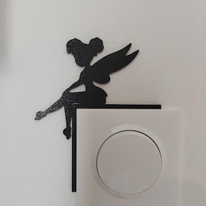 May include: Black metal fairy silhouette perched on a white electrical outlet. The fairy has detailed wings and is sitting with her legs dangling. The background is a plain white wall, creating a contrast with the dark figure.