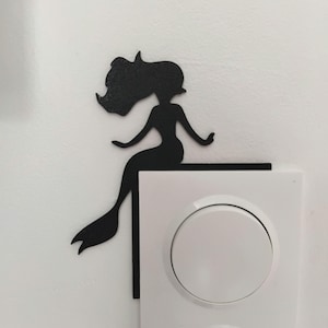May include: A black silhouette of a mermaid decorates a white electrical outlet. The mermaid is perched on the edge of the outlet, with flowing hair and a fishtail. The outlet has two white circular switches and a three-prong outlet.