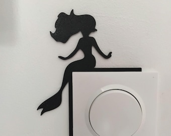 May include: A black mermaid silhouette sits on a white electrical outlet. The mermaid has flowing hair and a fishtail. The outlet features two white circular switches and a three-pin socket. The design adds a decorative touch.