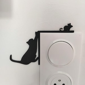 Könnte beinhalten: Schwarze Wanddekoration aus Metall mit Katze und Maus. Die Katze hängt an einer Kante, während die Maus oben sitzt. Die Dekoration befindet sich neben einem weißen Lichtschalter und einer Steckdose.