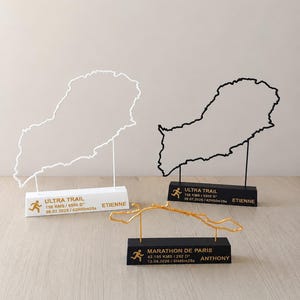 May include: Three running race trophies. Two feature outline maps, one white and one black, with the text "ULTRA TRAIL" and race details. The third is a gold wireframe of a race course, with "MARATHON DE PARIS" and race details.