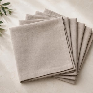 Può includere: Una pila di sei tovaglioli di stoffa quadrati color beige chiaro con una trama visibile. I tovaglioli sono piegati ordinatamente e sovrapposti l'uno sull'altro, mostrando un colore neutro uniforme e una finitura con orlo semplice.