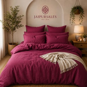 May include: A deep magenta bedding set featuring a duvet cover and four matching pillows. A cream-colored throw blanket with fringe edges is draped across the foot of the bed. The wall above the bed displays the text JAIPURSALES EST. 2018.