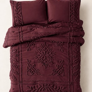 May include: A deep burgundy bedding set consisting of a comforter and two matching pillow shams. The fabric features a raised, tufted texture with intricate botanical and geometric patterns, including leaf motifs and a central starburst design.