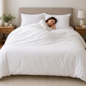 May include: A bed with white bedding, including a duvet cover and two pillows, is the focal point of the image. A person is sleeping on the bed. Two wooden nightstands flank the bed, each with a lamp and decorative items. The room has a neutral color palette.