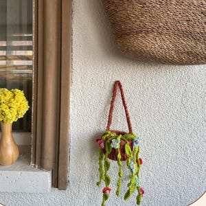 May include: A crocheted hanging plant in a brown pot with green vines and small multi-colored flowers. The plant features a brown crocheted handle for hanging. The flowers are light blue, pink, and red, attached to the green crocheted foliage.