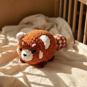 Pode incluir: Um brinquedo de pelúcia de panda vermelho feito à mão com detalhes brancos e uma cauda fofa repousa em um berço de madeira. Acima, um móbile com nuvens de feltro e contas de madeira está pendurado. O berço é preenchido com um cobertor bege macio.