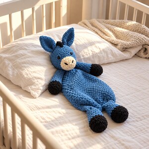 May include: A blue donkey-shaped crochet lovey featuring a cream-colored muzzle, black hooves, and black tufted hair. The plush toy has a textured, knotted yarn construction with two upright ears and small dark eyes, designed as a soft security blanket.