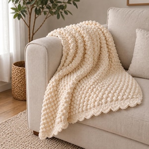 Può includere: Una coperta all'uncinetto color crema caratterizzata da una texture a rilievo con punto nocciolina su tutto il tessuto. La coperta presenta un bordo decorativo smerlato ed è drappeggiata sul bracciolo di un divano, evidenziando la sua struttura.