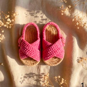 May include: A pair of bright pink crochet sandals featuring a crisscross strap design over the top of the foot. The sandals have a tan-colored crocheted sole and a small button detail on the side of each strap, showcasing a textured yarn construction.