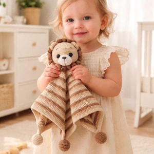 May include: A crochet lion security blanket in beige and brown. The lion's head has a detailed mane and a sweet face. The blanket features striped patterns and pom-poms at the corners. A soft, cuddly item for babies and toddlers, perfect for comfort.