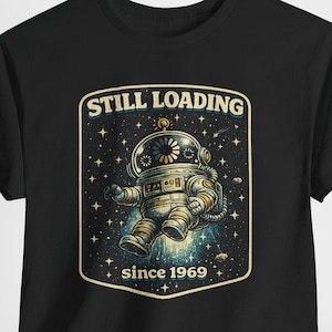 May include: Black t-shirt featuring a retro-style illustration of an astronaut in a spacesuit. The design includes the text "STILL LOADING" above the astronaut and "since 1969" below. The astronaut is set against a starry background.