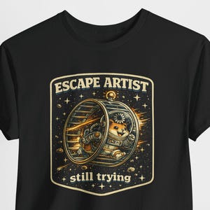 Può includere: T-shirt nera con un criceto dei cartoni animati in tuta spaziale dentro una ruota. La grafica include il testo "ESCAPE ARTIST" e "still trying". Il design utilizza colori oro, marrone e bianco. La t-shirt è di vestibilità standard.