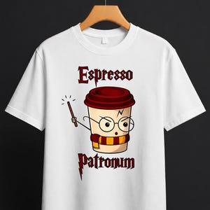 Può includere: T-shirt bianca con un disegno di tazza di caffè dei cartoni animati. La tazza indossa occhiali e una sciarpa, tenendo in mano una bacchetta. Le parole "Espresso Patronum" sono stampate sopra e sotto la tazza in un carattere stilizzato. Il design utilizza tonalità marroni, rosse e gialle.
