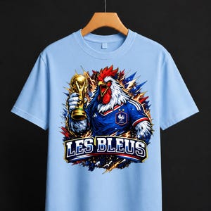 May include: Light blue t-shirt featuring a rooster wearing a French soccer jersey and holding a gold trophy. The shirt has the text "LES BLEUS" in a bold font. The rooster is depicted with red, white, and blue accents, symbolizing the French flag.