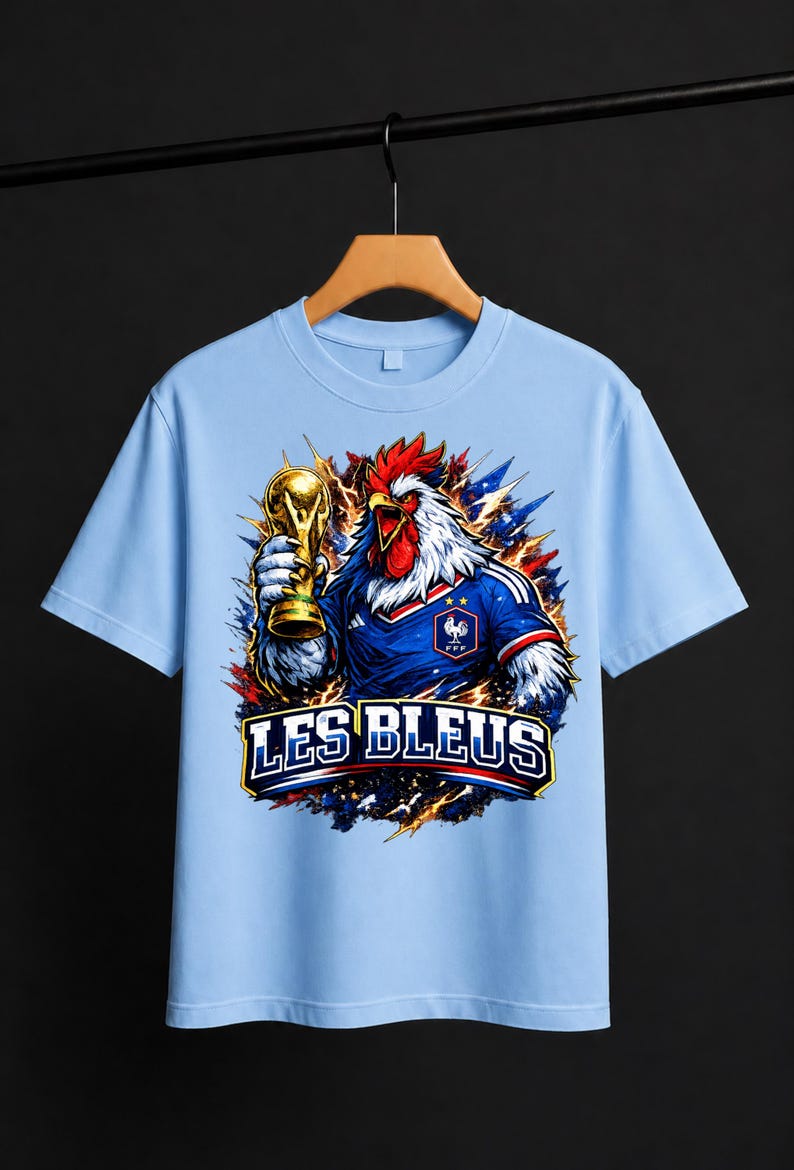 May include: Light blue t-shirt featuring a rooster wearing a French soccer jersey and holding a gold trophy. The shirt has the text "LES BLEUS" in a bold font. The rooster is depicted with red, white, and blue accents, symbolizing the French flag.