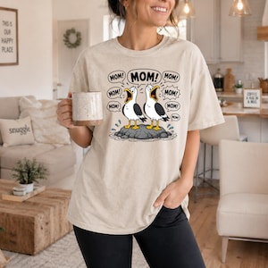 Può includere: T-shirt beige con un cartone animato di due gabbiani su una roccia, con fumetti che dicono "MOM!". La maglietta ha anche il testo "THIS IS OUR HAPPY PLACE" e un cuscino "snuggle".