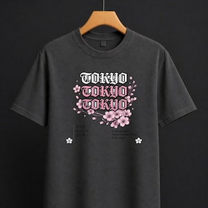 Può includere: T-shirt grigio scuro con la scritta "TOKYO" in bianco e rosa, insieme a grafiche di fiori di ciliegio. La maglietta presenta anche testo giapponese e la frase "A spring night, Cherry blossoms dance in the wind."
