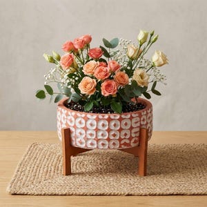 Puede incluir: Una maceta redonda de terracota con un diseño geométrico blanco, que contiene un ramo de rosas melocotón y crema, eucalipto y paniculata. La maceta está sobre un soporte de madera y un mantel individual de yute tejido.