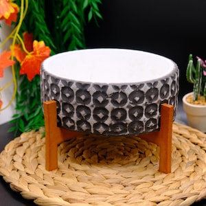 Könnte beinhalten: Ein runder, grau-weißer Pflanztopf mit geometrischem Muster, der von einem Holzständer getragen wird. Der Pflanztopf steht auf einer gewebten Matte, mit Grün im Hintergrund. Der Pflanztopf ist für den Innen- oder Außenbereich konzipiert.