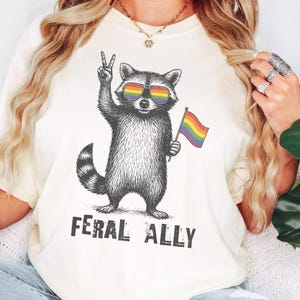 Pode incluir: Uma t-shirt de cor creme com uma ilustração a preto e branco de um guaxinim com óculos de sol arco-íris, fazendo o sinal de paz e segurando uma bandeira arco-íris, com o texto FERAL ALLY impresso por baixo.