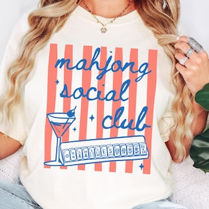 Könnte beinhalten: Cremefarbenes T-Shirt mit blau-roter Grafik. Das Design zeigt den Text "mahjong social club", einen Cocktail und eine Reihe von Mahjong-Steinen. Ein lässiges T-Shirt für Mahjong-Enthusiasten.