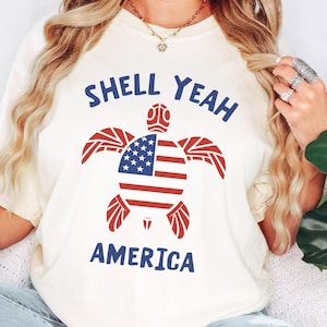 Könnte beinhalten: Cremefarbenes T-Shirt mit einem patriotischen Schildkröten-Design. Der Schildkrötenpanzer zeigt die amerikanische Flagge mit roten und weißen Streifen und einem blauen Bereich mit weißen Sternen. Der Text "SHELL YEAH AMERICA" ist über und unter der Schildkröte in Blau gedruckt.