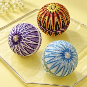 Puede incluir: Tres bolas temari esféricas con intrincados patrones de bordado geométrico. Una bola es morada y blanca, otra es negra con detalles en amarillo y naranja, y otra es blanca con detalles en hilo azul y verde, todas con un bordado preciso.