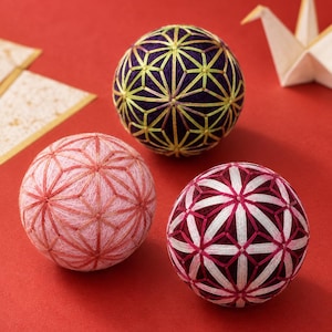May include: Three spherical temari balls featuring intricate geometric thread embroidery. One ball is deep purple with yellow stitching, one is light pink with matching pink thread, and one is dark maroon with white and pink star-shaped patterns.