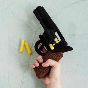 May include: A handmade crochet toy gun in black and brown, with yellow accents. The toy gun is held in a hand, and two yellow crochet bullets are next to the gun. The toy gun is a unique, handcrafted item.