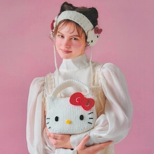 Könnte beinhalten: Eine weiße gehäkelte Handtasche in Form von Hello Kitty mit roter Schleife, schwarzen Augen und gelber Nase. Das Set enthält ein passendes weißes gehäkeltes Stirnband mit Hello Kitty-Gesichtern an den Seiten und kleinen roten Schleifen.