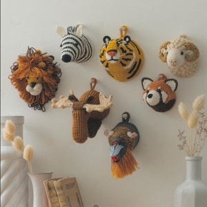 Puede incluir: Una colección de siete cabezas de animales de ganchillo montadas en la pared, que incluye un león con melena de hilo, una cebra blanca y negra, un tigre amarillo y negro, un carnero color crema, un alce marrón con astas, un panda rojo y un mandril.