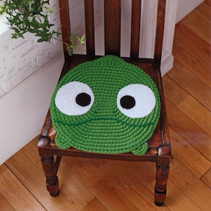 May include: A round, green crocheted cushion featuring two large white circular patches with black centers to resemble eyes. The cushion has a simple dark green stitched line for a mouth and small protruding shapes at the bottom edge.
