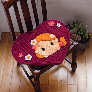 May include: A heart-shaped crochet cushion in deep magenta featuring a central applique of a cartoon character with orange hair and a peach-colored face. The cushion is decorated with several small, multi-colored crocheted flowers.