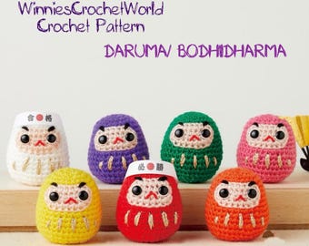 Könnte beinhalten: Eine Sammlung von sieben gehäkelten Daruma-Puppen in verschiedenen Farben, darunter Weiß, Lila, Grün, Rosa, Gelb, Rot und Orange. Jede Puppe hat ein unverwechselbares Gesicht und ist ein handgefertigtes Häkelmuster.